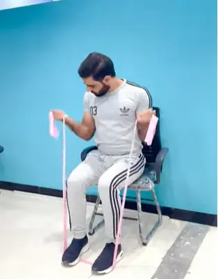 A man performs a seated biceps curl using a resistance band while sitting on a chair. He holds the band with both hands, doing Biceps Workout at Home, engaging his biceps as he curls his arms upward.